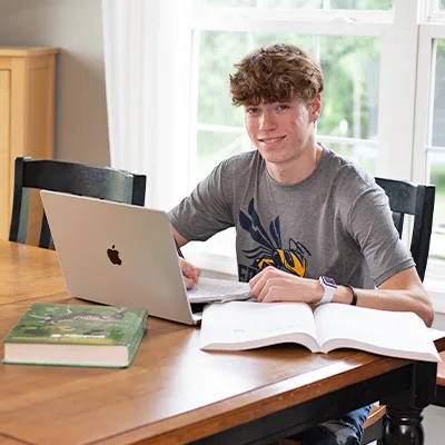 Sultan-Dual-Enrollment-studying Male high school student smiling while sitting at table with laptop and books.