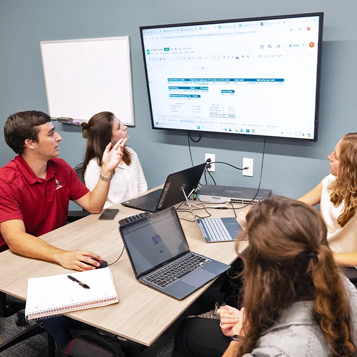 Four students gathered at table with laptops and notebooks looking at a spreadsheet on TV screen.