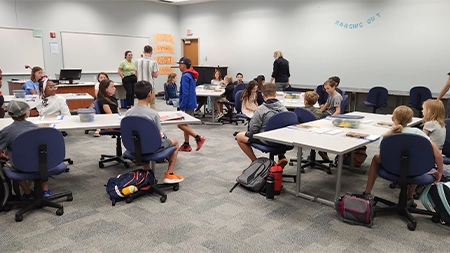 Middle school students sitting around tables in classroom.