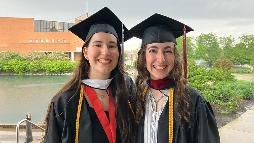 Emily Vest and Brianna De Man celebrate their graduation from Cedarville University in their caps and gowns with Cedar Lake and the Dixon Ministry Center in the background