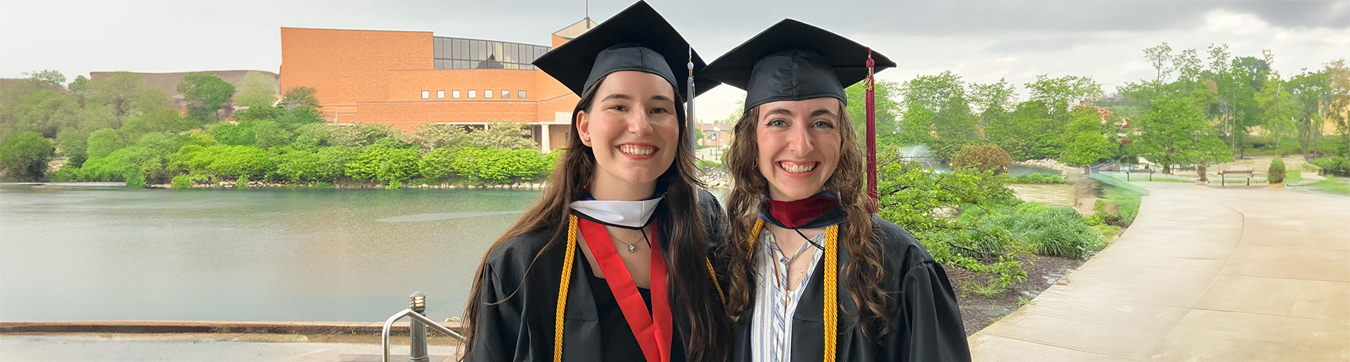 Emily Vest and Brianna De Man celebrate their graduation from Cedarville University in their caps and gowns with Cedar Lake and the Dixon Ministry Center in the background