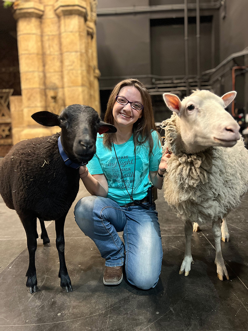 Abigail King on theatre stage smiling next to two sheep.