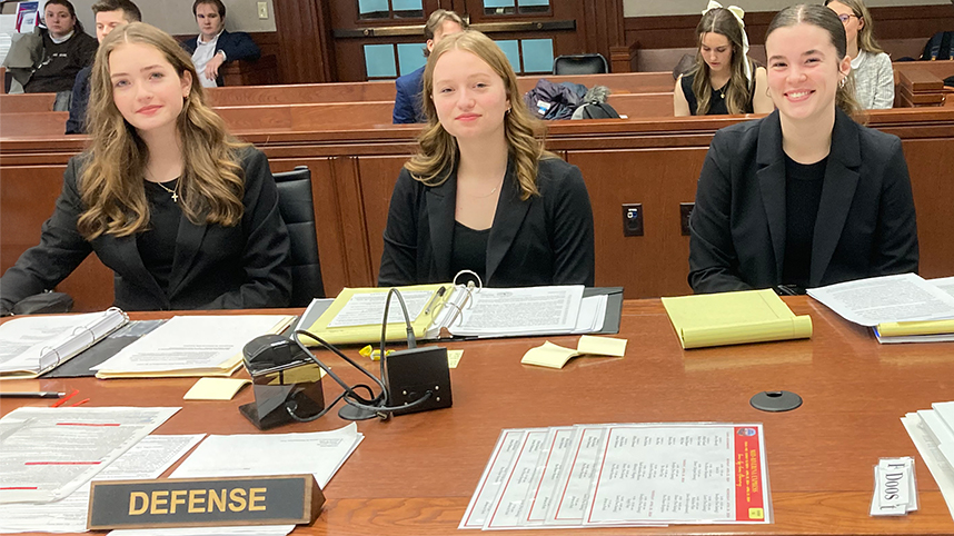 Three female students in professional clothes smiling behind courtroom desk with papers.