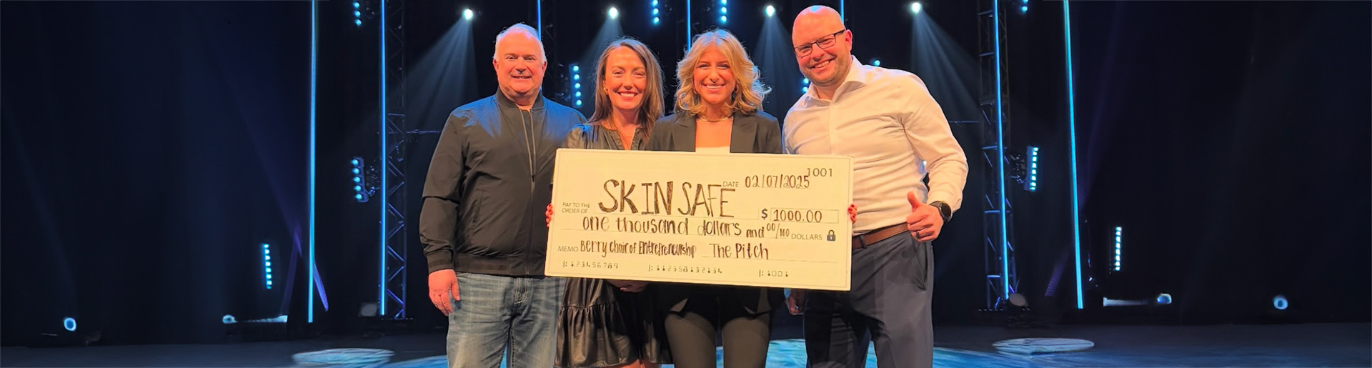 Alex Simons smiling onstage with three judges and holding large check.