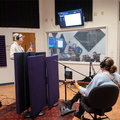 Students record music in a campus audio studio while classmates monitor the session from the control room.