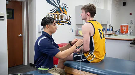 An athletic trainer applies electrical stimulation therapy to a basketball player’s arm in a university training room.