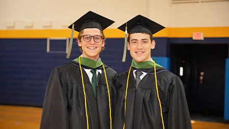 Two graduates smiling in graduation caps and gowns in the gym.