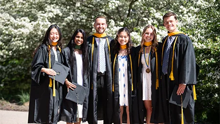 Six graduates smiling in caps and gowns outside by flowering trees.