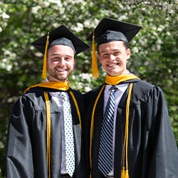 Two male graduates in caps and gowns smiling with arms around each other in front of flowering bushes.