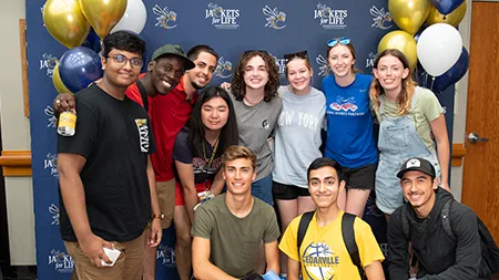 Group of eleven students smiling in front of Jackets for Life photo backdrop.