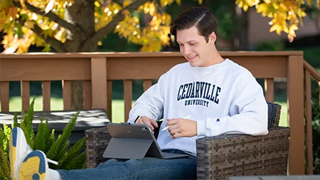 Student sitting in chair on porch studying and holding a tablet.