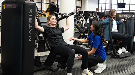 A fitness trainer assists a woman using a chest press machine in a gym, with other people exercising in the background.