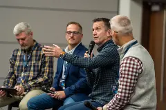 Four men seated in chairs, engaged in discussion while speaking into microphones during a conference session.