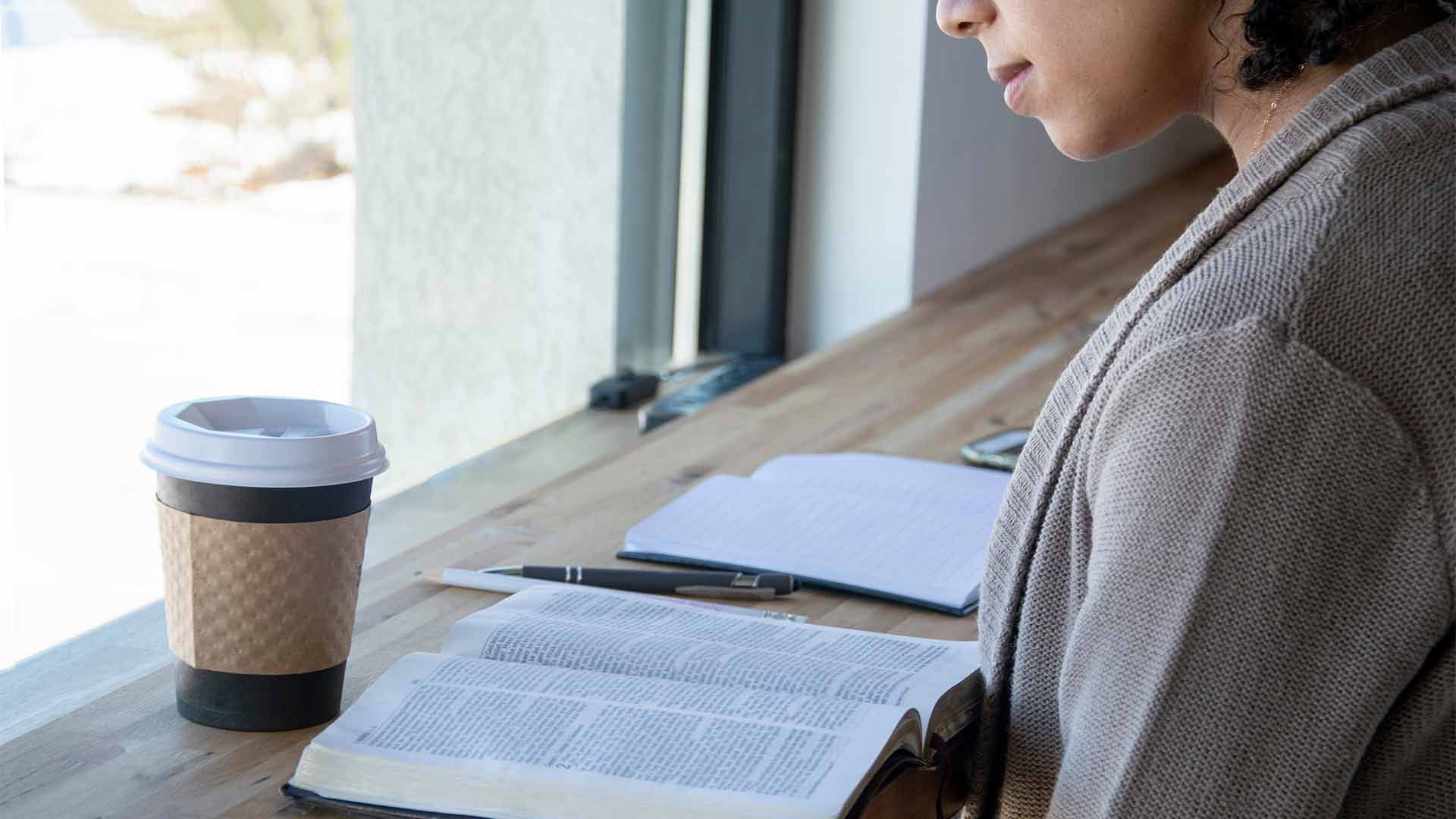 Woman reading her Bible at long table with coffee cup.