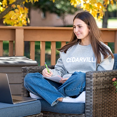 Student sitting outside on a patio chair, wearing a Cedarville sweatshirt, taking notes while looking at a laptop.