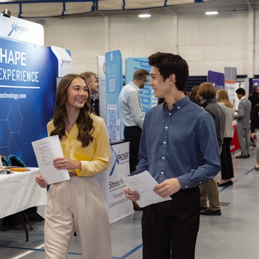 Two students at a career fair, smiling and holding resumes while walking past booths and interacting with other attendees.