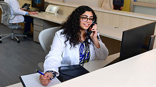 Female Pharmacy student dressed in white lab coat at a desk making phone calls to help with contact tracing.
