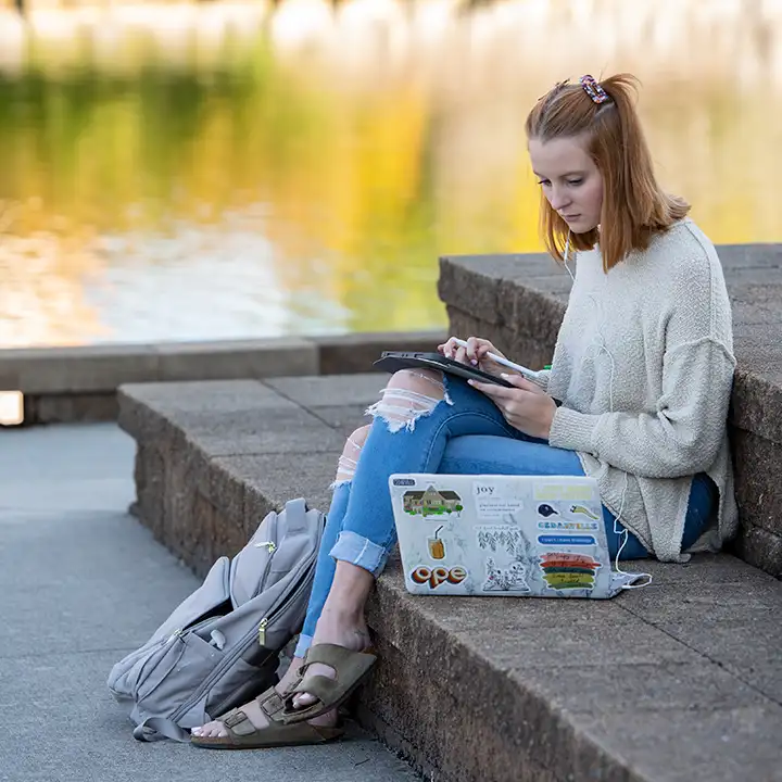 Woman sitting on outside steps by a pond working on laptop.